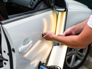 Technician repairing dented cars door using paintless dent removal method under bright light reflection.